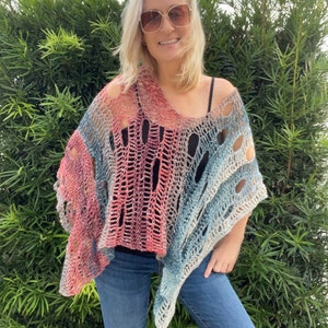 May include: A handmade, open-weave poncho in shades of pink, blue, and silver. The poncho has a loose, draped fit and is made of a soft, textured yarn. The garment is worn over a black top and blue jeans.