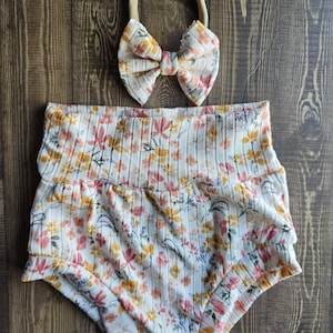May include: A baby outfit set featuring a floral print. The set includes high-waisted bloomers and a matching bow headband. The fabric has a ribbed texture and a pattern of yellow, pink, and blue flowers on a white background.