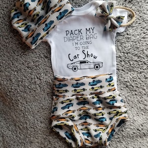 Könnte beinhalten: Ein weißer Baby-Strampler mit dem Text "Pack My Diaper Bag I'm Going To The Car Show" und einem Comic-Auto. Der Strampler wird mit einem weißen Stirnband mit einer blauen und gelben Schleife, einer weißen Mütze mit einem blauen und gelben Auto-Muster und weißen Blümchen mit einem blauen und gelben Auto-Muster kombiniert.