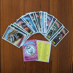 May include: A collection of 25 Skeleton Pile Kids trading cards featuring various cartoon skeletons. The cards are in a variety of colors, including black, white, red, and blue. The cards are arranged in a fan shape, with the checklist card at the bottom. The checklist card has a pink background and features a cartoon skeleton with a skull and crossbones.