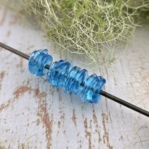 May include: A close-up of a series of translucent blue glass beads with a spiral design strung on a dark metal rod. The beads are a vibrant shade of blue. The background features a blurred, textured surface and green foliage.
