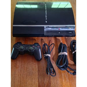 May include: A black Sony PlayStation 3 game console with a glossy finish, featuring the text PlayStation 3 on the top. Included is a black wireless controller with Sony branding and three black power and connection cables bundled with rubber bands.