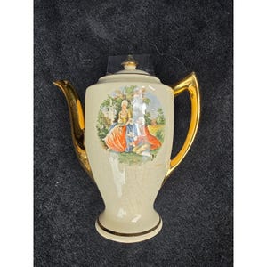 May include: A cream-colored ceramic teapot featuring a central circular illustration of two figures in period clothing within a garden setting. The teapot has a gold-toned handle, spout, and lid finial, with a fine crackle glaze finish throughout.