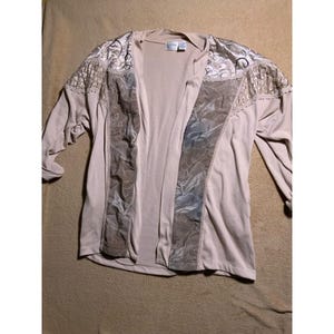 May include: A beige open cardigan with long sleeves. The cardigan features decorative panels with a floral pattern in shades of gray and beige. The shoulders have a textured, patterned design. The garment is made of a soft, lightweight material.