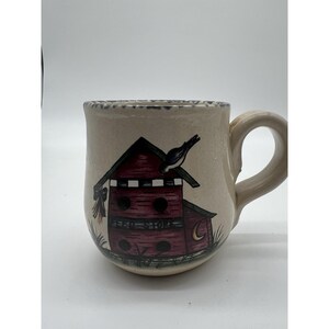 May include: Cream-colored ceramic mug with a birdhouse design. The birdhouse is red with a black bird perched on top. The handle is a matching cream color, and the rim has a blue and white pattern.