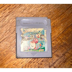May include: A gray Nintendo Game Boy video game cartridge for Kirby's Dream Land 2. The rectangular plastic casing features a worn, partially peeled label displaying the game title, Nintendo branding, and colorful character illustrations.