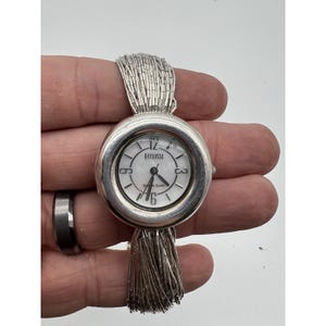 Puede incluir: Un reloj Eclissi plateado con esfera blanca y correa de cuentas plateadas. La esfera del reloj tiene números y manecillas negras. La correa está hecha de pequeñas cuentas plateadas. El reloj se sostiene en una mano.