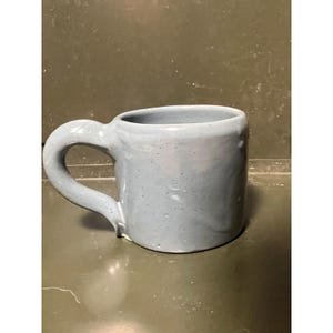 May include: A light blue ceramic mug with a glossy, speckled finish and a thick, rounded handle. The mug features an irregular, hand-formed shape with a smooth texture and a wide opening, showcasing a consistent pale blue color throughout the body.