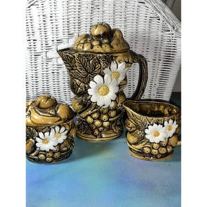 May include: A vintage ceramic tea set in shades of brown and gold, featuring a teapot with a lid, a sugar bowl, and a creamer. Each piece is adorned with white daisy accents and grape cluster designs.