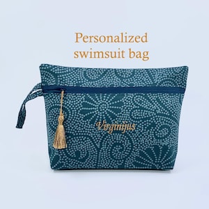 May include: Teal and white patterned swimsuit bag with a gold tassel zipper pull. The bag has a dark blue zipper and a handle. The text "Personalized swimsuit bag" is above the bag, and the name "Virginijus" is embroidered on the bag.
