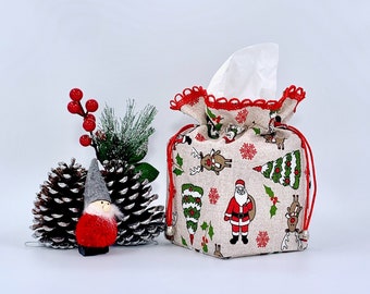 May include: A festive tissue box cover with a drawstring closure. The beige fabric is patterned with Christmas trees, Santa Claus, and reindeer. The top edge is trimmed with red scalloped fabric. A small gnome and pinecones are next to the tissue box.