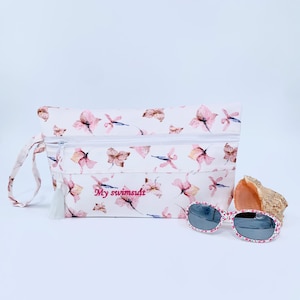 May include: A white zippered pouch with a floral print and the text "My swimsuit" embroidered on the front. The pouch has a white tassel and is next to a pair of pink and white sunglasses and a seashell.