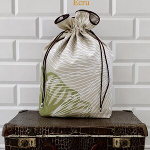 May include: An ecru drawstring bag with a white and green leaf pattern, brown drawstring, and a brown trim. The bag is sitting on a vintage brown suitcase. The word "Ecru" is in the top center.