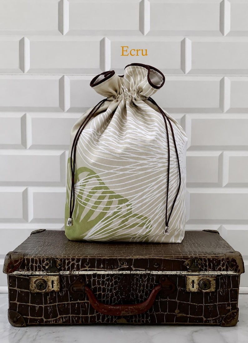 May include: An ecru drawstring bag with a white and green leaf pattern, brown drawstring, and a brown trim. The bag is sitting on a vintage brown suitcase. The word "Ecru" is in the top center.