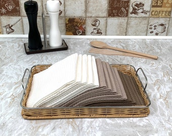 May include: A wicker basket with a silver handle holds a stack of white and brown kitchen towels. The basket is on a light gray countertop.