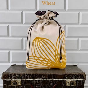 May include: A beige drawstring bag with a yellow floral design, black drawstrings, and silver accents. The bag is sitting on a vintage brown suitcase. The word "Wheat" is printed above the bag.