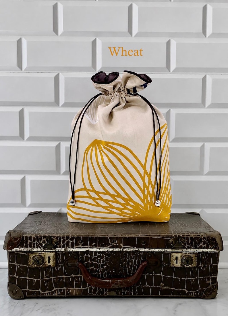 May include: A beige drawstring bag with a yellow floral design, black drawstrings, and silver accents. The bag is sitting on a vintage brown suitcase. The word "Wheat" is printed above the bag.