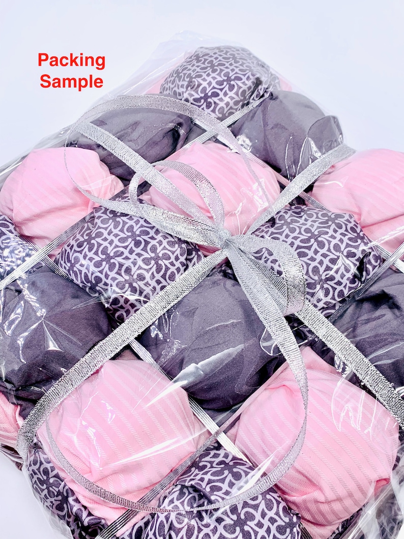 Op de afbeelding: Een doorzichtige plastic zak gevuld met roze en paarse vierkante stoffen, vastgebonden met een zilveren lint. De zak is gelabeld met "Packing Sample".