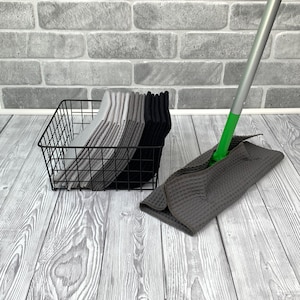 Puede incluir: Una cesta de alambre negra llena de toallas de cocina grises y negras. Una mopa gris y verde con un mango largo está tumbada en un suelo de madera gris claro.