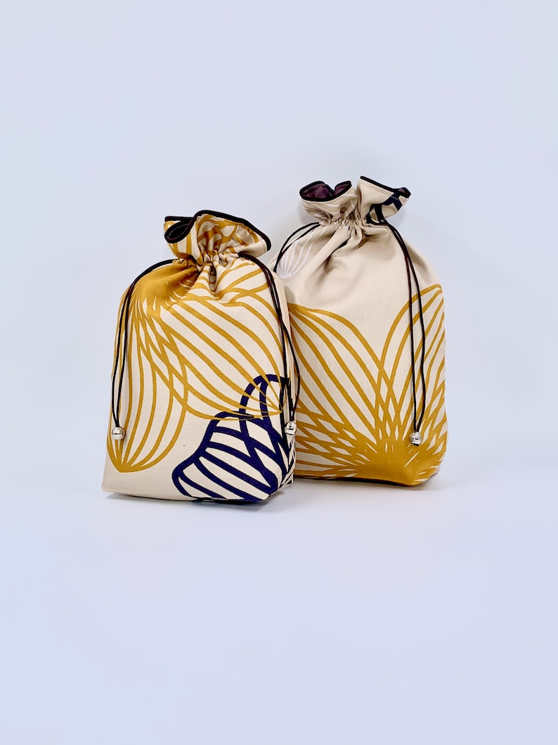May include: Two drawstring bags with a cream background and a gold and navy blue leaf pattern. The bags have black drawstrings with silver-colored beads and black trim. The bags are tied at the top.