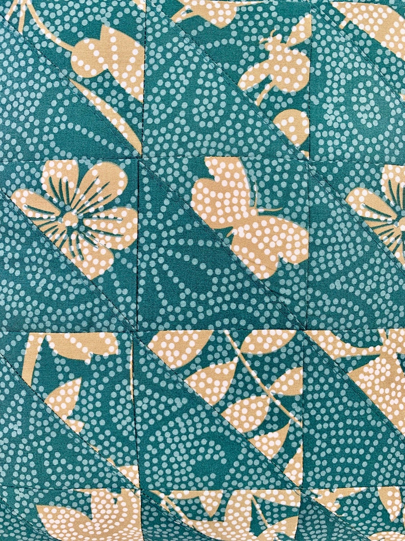 以下が含まれることがあります： 白のドット柄とベージュの花柄が施されたターコイズブルーの生地。生地は三角形にカットされ、パッチワークパターンになるように縫い合わされています。