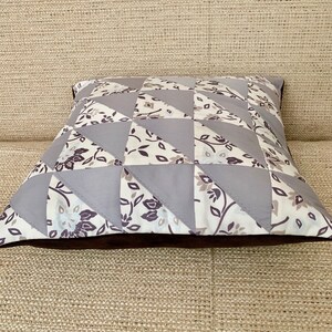 May include: A square cushion with a patchwork design featuring grey and white fabric with floral patterns. The cushion has a brown border.