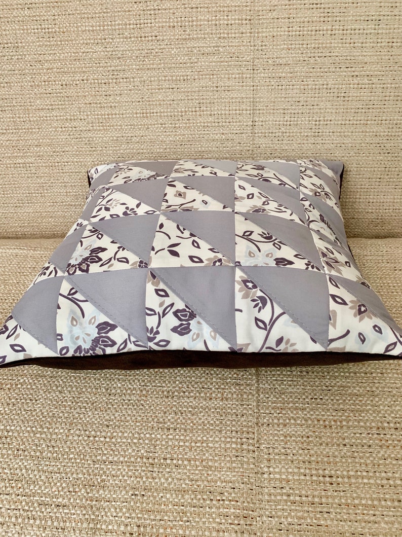 May include: A square cushion with a patchwork design featuring grey and white fabric with floral patterns. The cushion has a brown border.