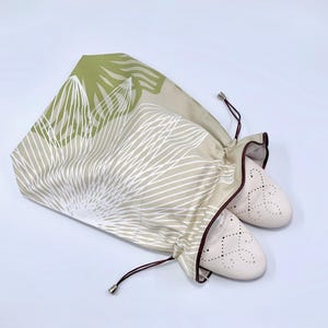May include: A pair of beige ballet flats peeking out of a drawstring shoe bag. The bag is patterned with white linear designs and green leaf shapes on a tan background. The bag has a brown trim and drawstring.