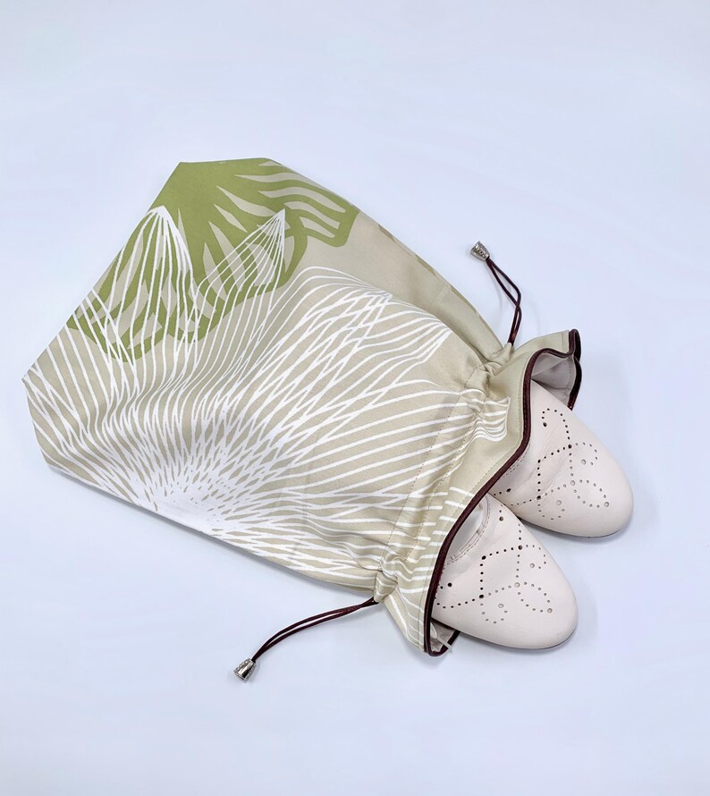 May include: A pair of beige ballet flats peeking out of a drawstring shoe bag. The bag is patterned with white linear designs and green leaf shapes on a tan background. The bag has a brown trim and drawstring.