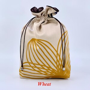 May include: A beige drawstring bag with a large yellow floral design. The bag has a brown drawstring and trim. The word "Wheat" is printed in red at the bottom of the bag.