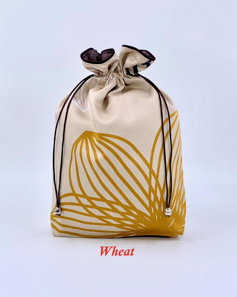May include: A beige drawstring bag with a large yellow floral design. The bag has a brown drawstring and trim. The word "Wheat" is printed in red at the bottom of the bag.