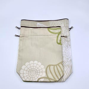 May include: Two drawstring bags in a neutral color with floral patterns. The bags have a beige background with white, green, and brown floral designs. The top edges are trimmed with a dark brown border and silver-toned metal eyelets.