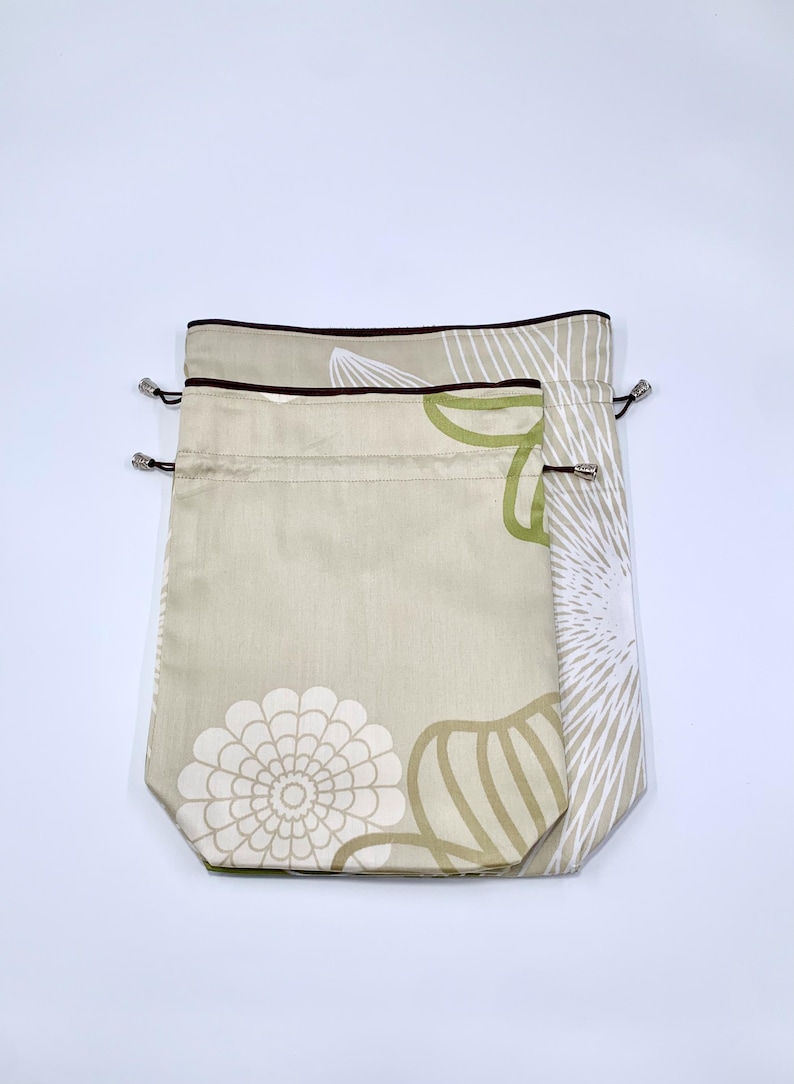 May include: Two drawstring bags in a neutral color with floral patterns. The bags have a beige background with white, green, and brown floral designs. The top edges are trimmed with a dark brown border and silver-toned metal eyelets.