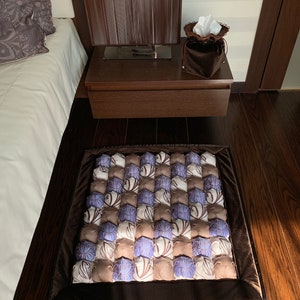 Op de afbeelding: Een bruin en wit gepatroneerd vloerkleed met een donkerbruine rand. Het kleed is gemaakt van afzonderlijke vierkanten, elk met een ander patroon. Het kleed ligt op een houten vloer.