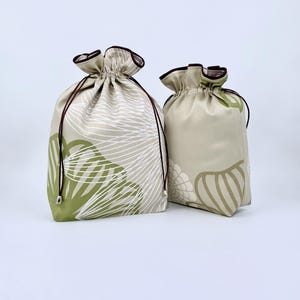 May include: Two drawstring bags with floral patterns. The larger bag features a white and green leaf design on a beige background. The smaller bag has a similar pattern in green and beige. Both bags have brown drawstrings and trim.