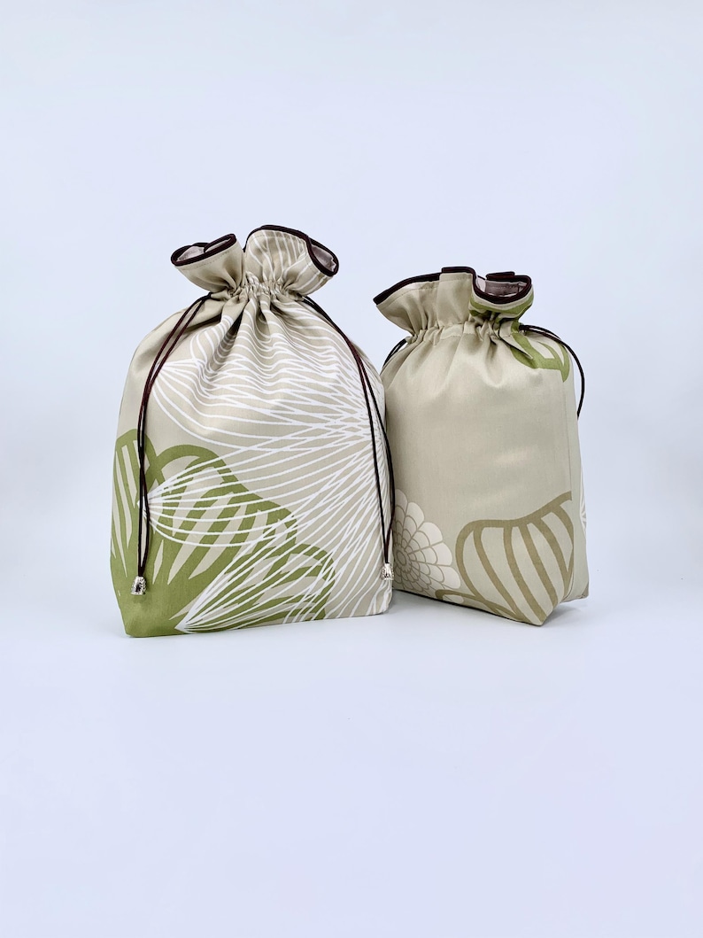 May include: Two drawstring bags with floral patterns. The larger bag features a white and green leaf design on a beige background. The smaller bag has a similar pattern in green and beige. Both bags have brown drawstrings and trim.