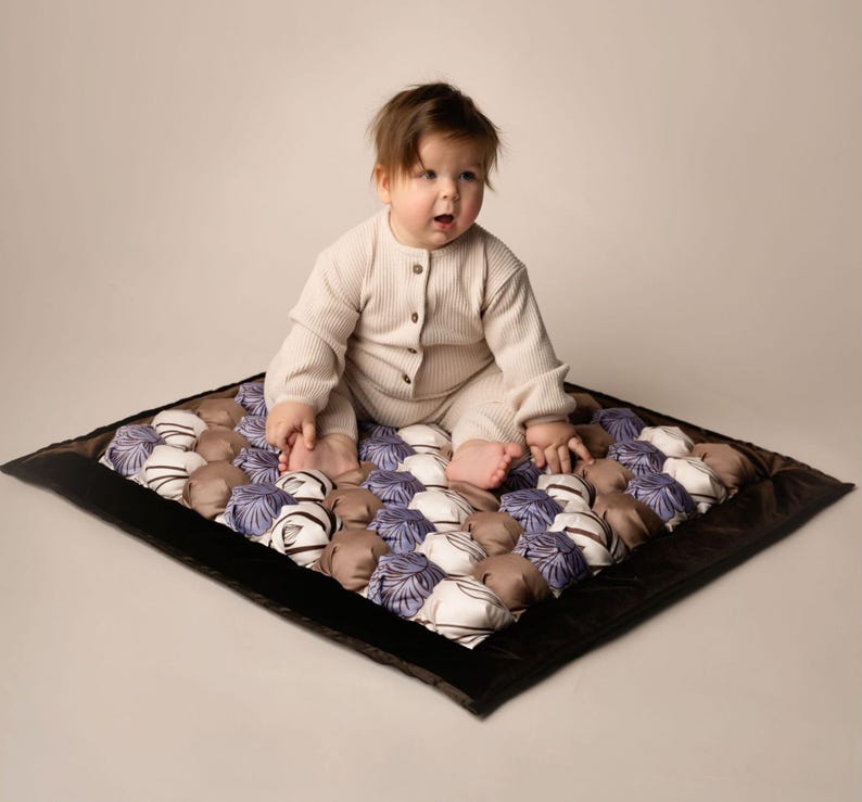 Op de afbeelding: Een baby in een cr&egrave;mekleurige romper zit op een veelkleurige, gevoerde speelmat met een bruine rand. De mat is gemaakt van vierkante stoffen met een gestikt ontwerp.