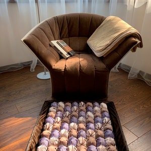 Op de afbeelding: Een bruine fauteuil met een beige deken en een boek met de titel "ABSOLUT ART" op de zitting. Voor de stoel ligt een bruin en paars, gepatroneerd vloerkleed met een bruine rand.
