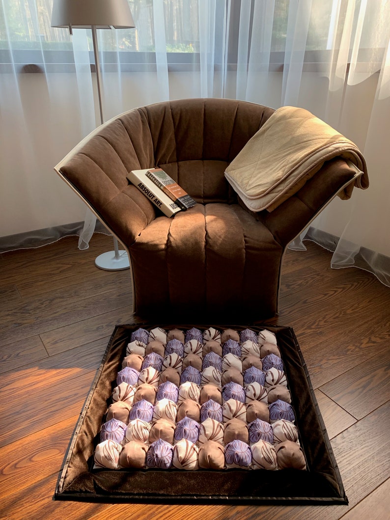 Op de afbeelding: Een bruine fauteuil met een beige deken en een boek met de titel "ABSOLUT ART" op de zitting. Voor de stoel ligt een bruin en paars, gepatroneerd vloerkleed met een bruine rand.