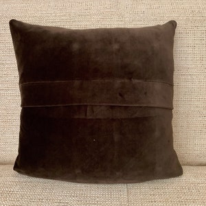 May include: A brown velvet cushion with a decorative seam down the centre. The cushion is sitting on a beige textured surface.