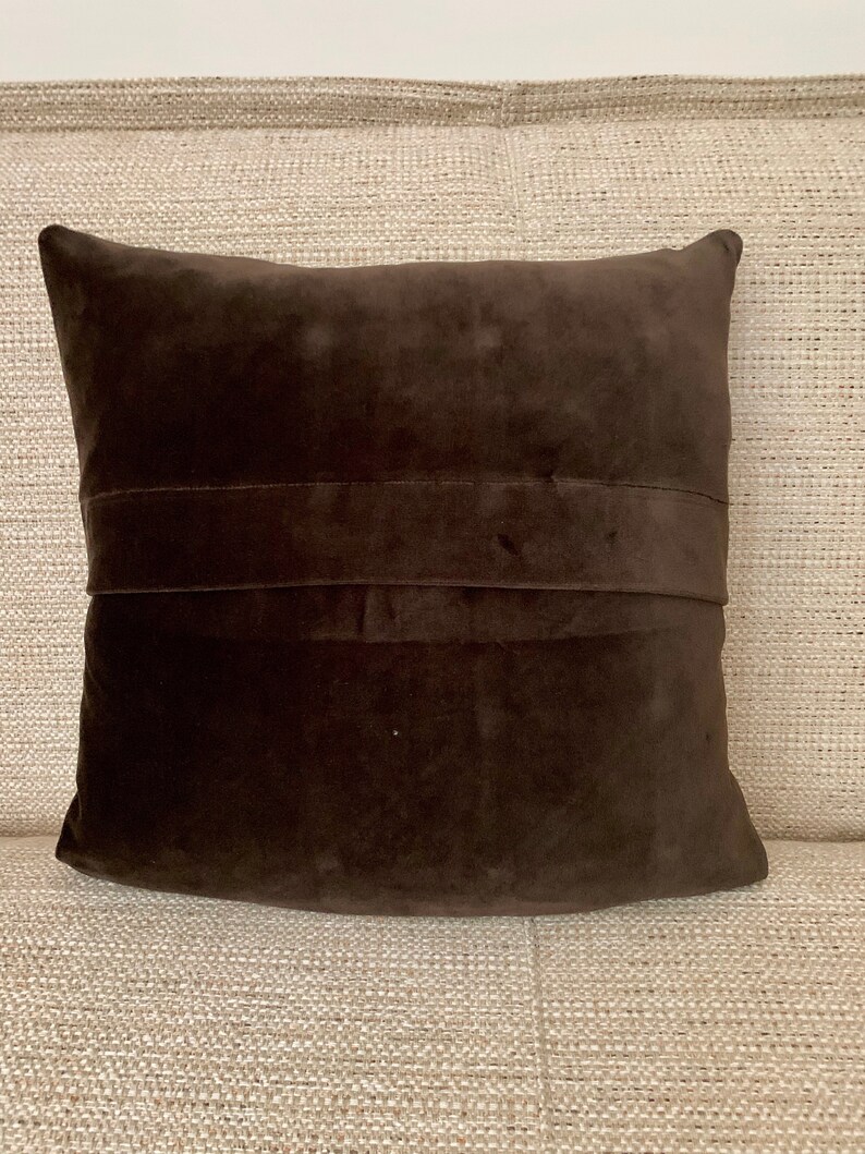 May include: A brown velvet cushion with a decorative seam down the centre. The cushion is sitting on a beige textured surface.