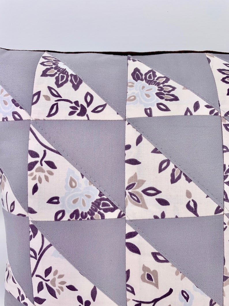 May include: A grey and white patterned cushion with a geometric design. The cushion is made of fabric with a floral pattern and is sewn together in a patchwork style.