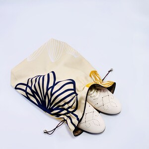 May include: Beige drawstring shoe bag with a navy and gold floral design. The bag is open, revealing a pair of white leather shoes with decorative cutouts. The bag's design features a repeating pattern of stylized leaves.
