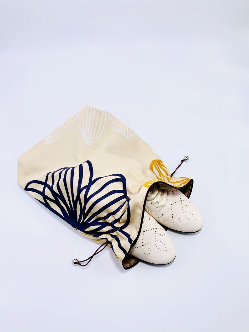 May include: Beige drawstring shoe bag with a navy and gold floral design. The bag is open, revealing a pair of white leather shoes with decorative cutouts. The bag's design features a repeating pattern of stylized leaves.