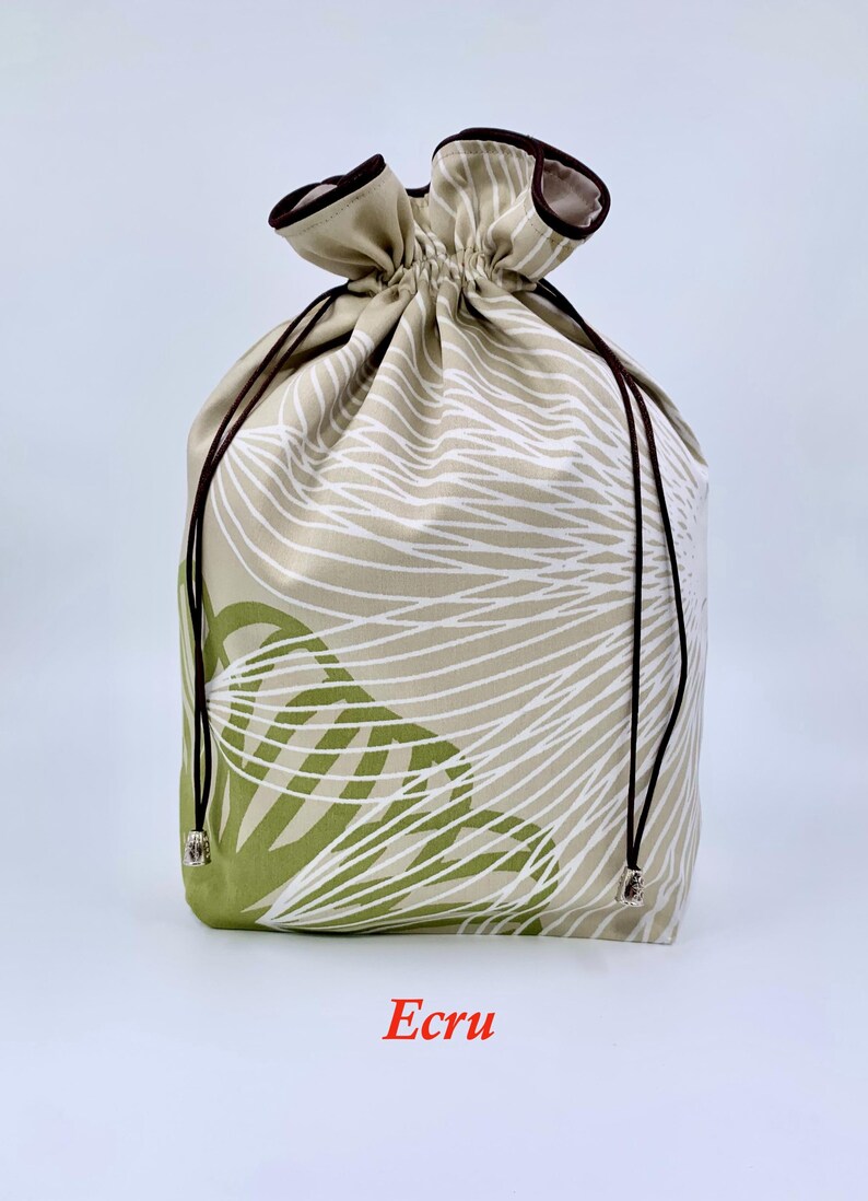 May include: An ecru-colored drawstring bag with a botanical pattern in white and green. The bag has a brown drawstring and trim. The word "Ecru" is printed at the bottom of the bag.