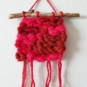 May include: A small, woven wall hanging in shades of red, pink, and burgundy. The textile art piece is suspended from a natural wooden branch with red yarn. The bottom edge features several dangling red yarn tassels.