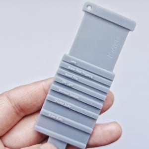 Pode incluir: Uma ferramenta cinza para medir fios com a palavra "Yarn" impressa. A ferramenta possui uma série de ranhuras rotuladas para medir o peso do fio, incluindo "Lace", "Super Fine" e "Super Bulky".