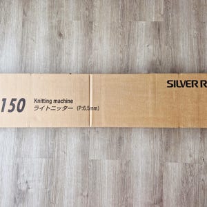 Può includere: Scatola rettangolare di cartone per una macchina per maglieria Silver Reed LK150. La scatola marrone chiaro presenta testo nero: "LK150 Knitting machine ライトニッター (P:6.5mm)" e "SILVER REED".