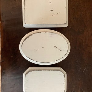 May include: Three off-white, distressed trays in various shapes: rectangular, oval, and octagonal. The trays have a slightly weathered look with visible wood grain and dark edges, set on a dark wood surface.