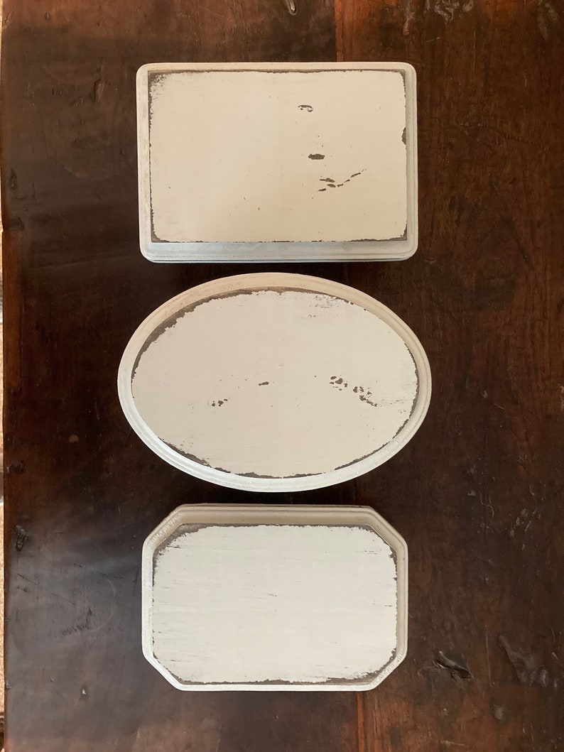 May include: Three off-white, distressed trays in various shapes: rectangular, oval, and octagonal. The trays have a slightly weathered look with visible wood grain and dark edges, set on a dark wood surface.